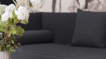 Ett rum med en mörkgrå soffa, en randig vas med vita blommor, en svartvit mugg och en tidning på ett träbord.