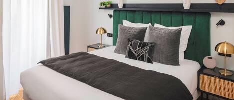 A modern bedroom with a green upholstered headboard, a round mirror, bedside lamps, and a bedside table with a speaker.