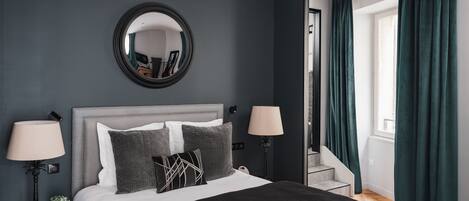 A modern bedroom with a grey headboard, a large bed with white and grey bedding, two bedside lamps, a round mirror above the bed, and a window with teal curtains.