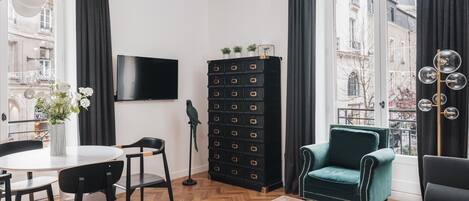 A modern living room with a wooden herringbone floor, a black storage unit, a green armchair, a black sofa, and a dining area with a white table and black chairs.