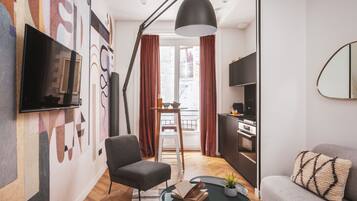 Un salon moderne avec un téléviseur à écran plat, un canapé, une table basse sur laquelle se trouvent un livre et une plante, un grand lampadaire et une vue sur la ville par la fenêtre.