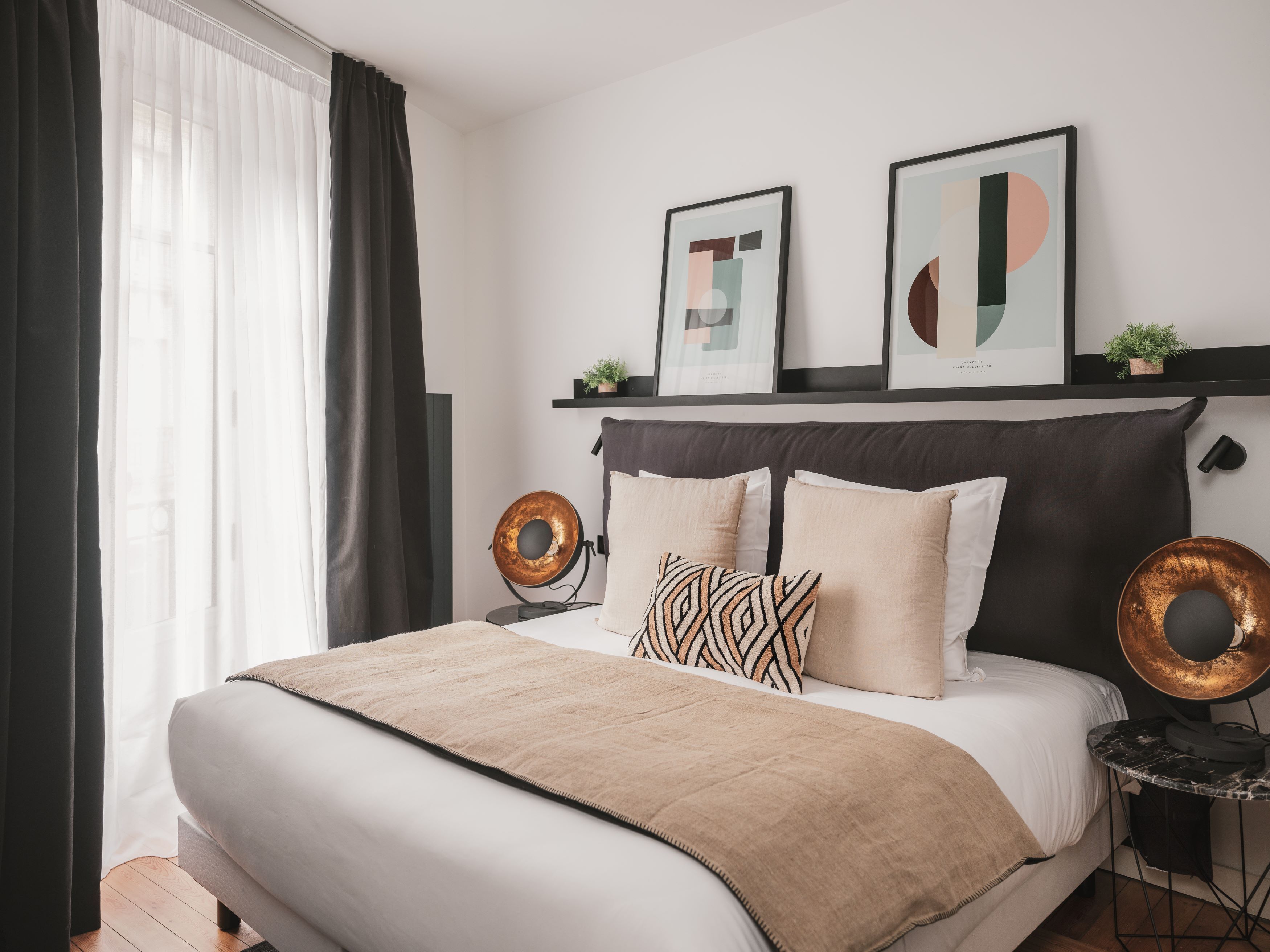 Un dormitorio moderno con una cama grande, un cabecero, dos obras de arte abstractas enmarcadas, una mesita de noche con una lámpara y una ventana con cortinas de gasa.