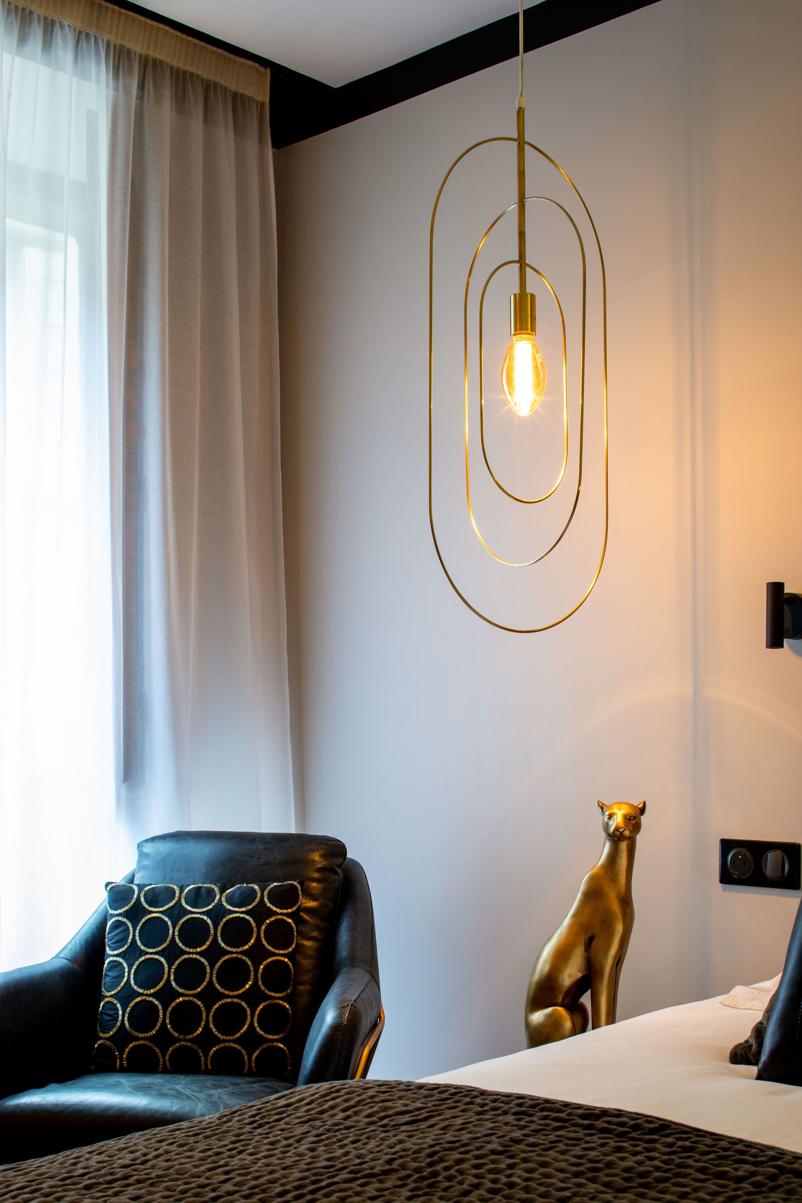 A modern hotel room with a leather armchair, a decorative pillow, a wall-mounted light fixture, and a golden statue.