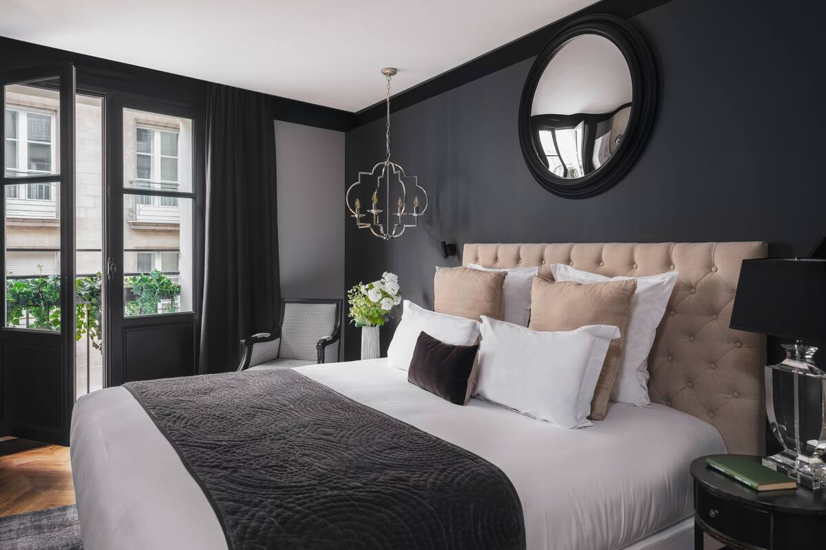 A modern hotel room with a large bed, a round mirror, a chandelier, and a view of the city through large windows.