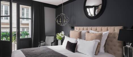 A modern hotel room with a large bed, a round mirror, a chandelier, and a view of the city through large windows.