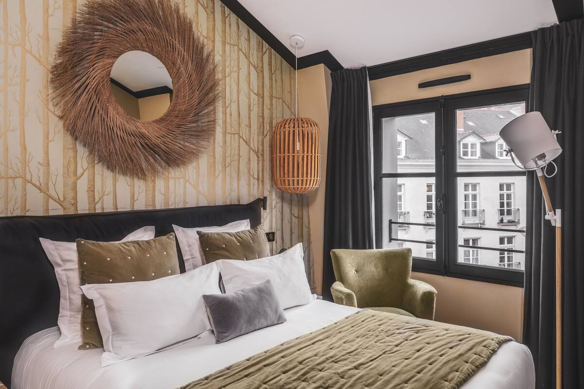 A hotel room with a bed, a green armchair, a round mirror, and a window with a view of buildings.