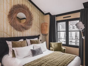 Ein Hotelzimmer mit einem Bett, einem grünen Sessel, einem runden Spiegel und einem Fenster mit Blick auf Gebäude.