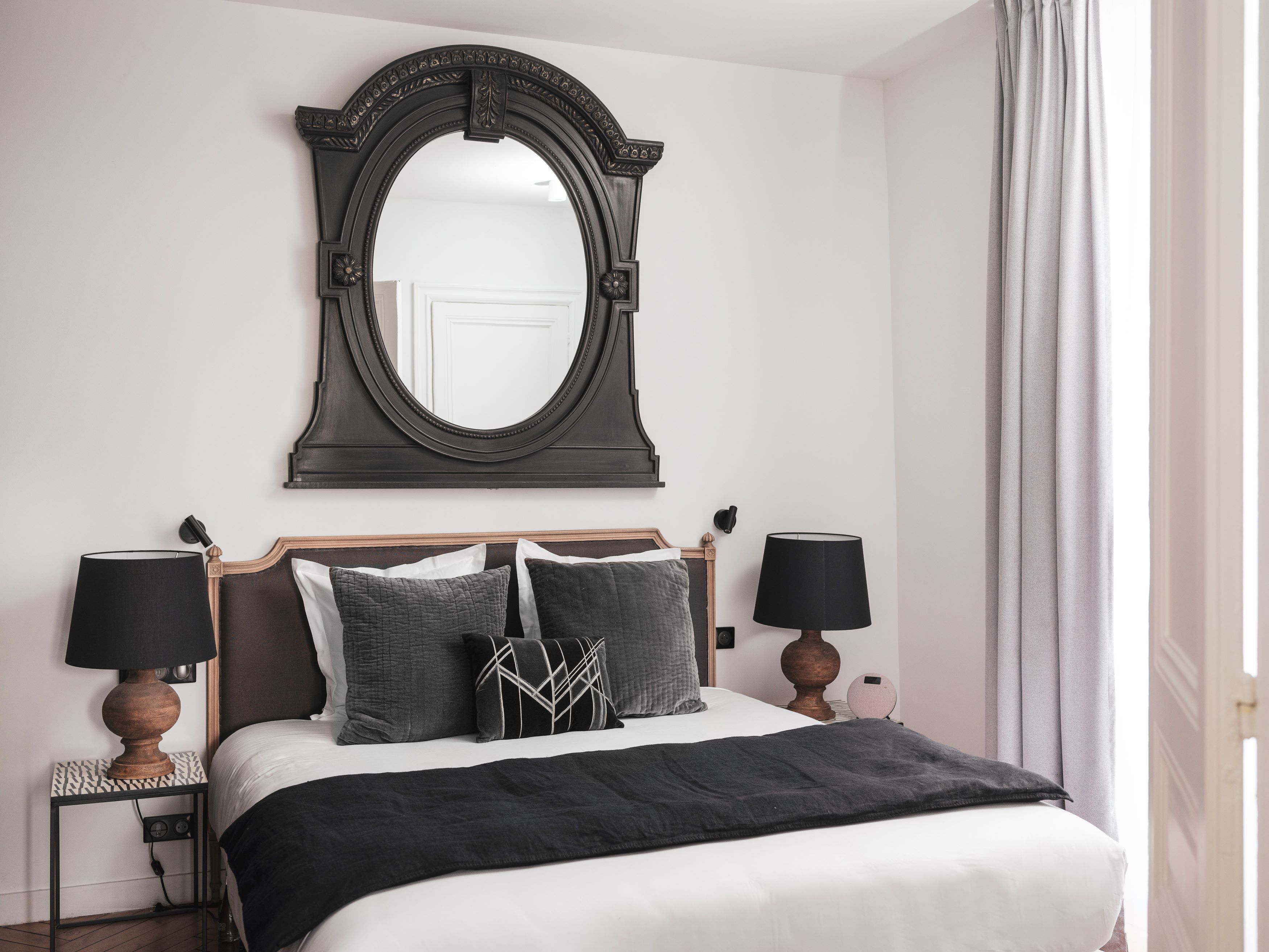 Une chambre à coucher bien rangée, avec un grand miroir, deux lampes de chevet, un lit recouvert d’une couvre-lit noir et blanc, et une table de chevet avec une lampe.