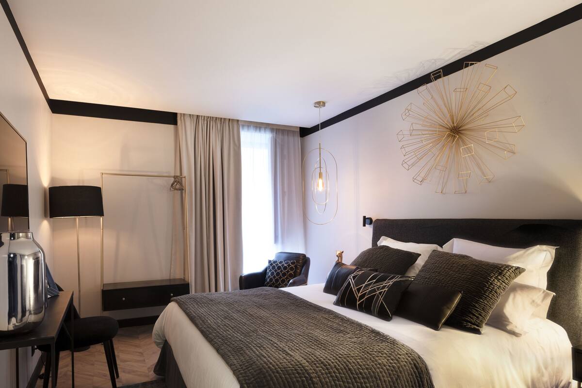 A modern hotel room with a large bed, a desk with a coffee machine, a chair, and a large mirror.