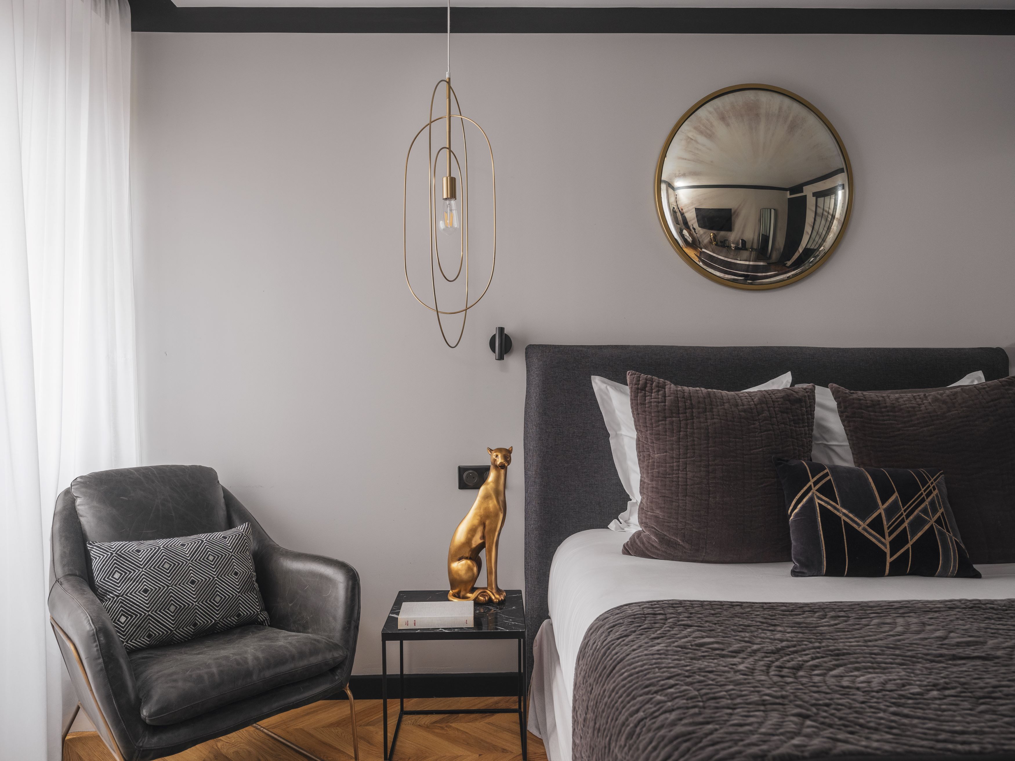 A modern bedroom with a dark grey sofa, a round mirror, a gold statue, and a bed with multiple pillows.