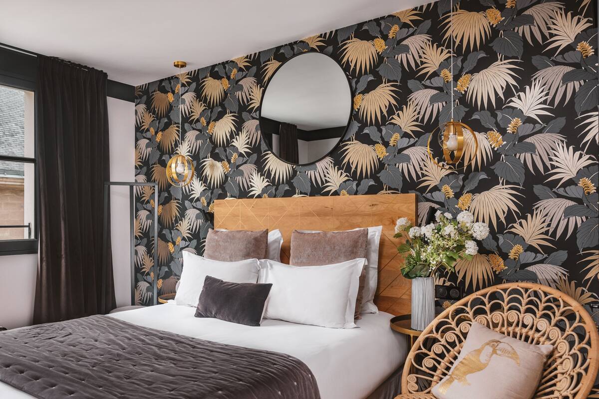 A bedroom with a bed, dark curtains, a round mirror, a wall with a tropical leaf pattern, and a wicker chair with a cushion.