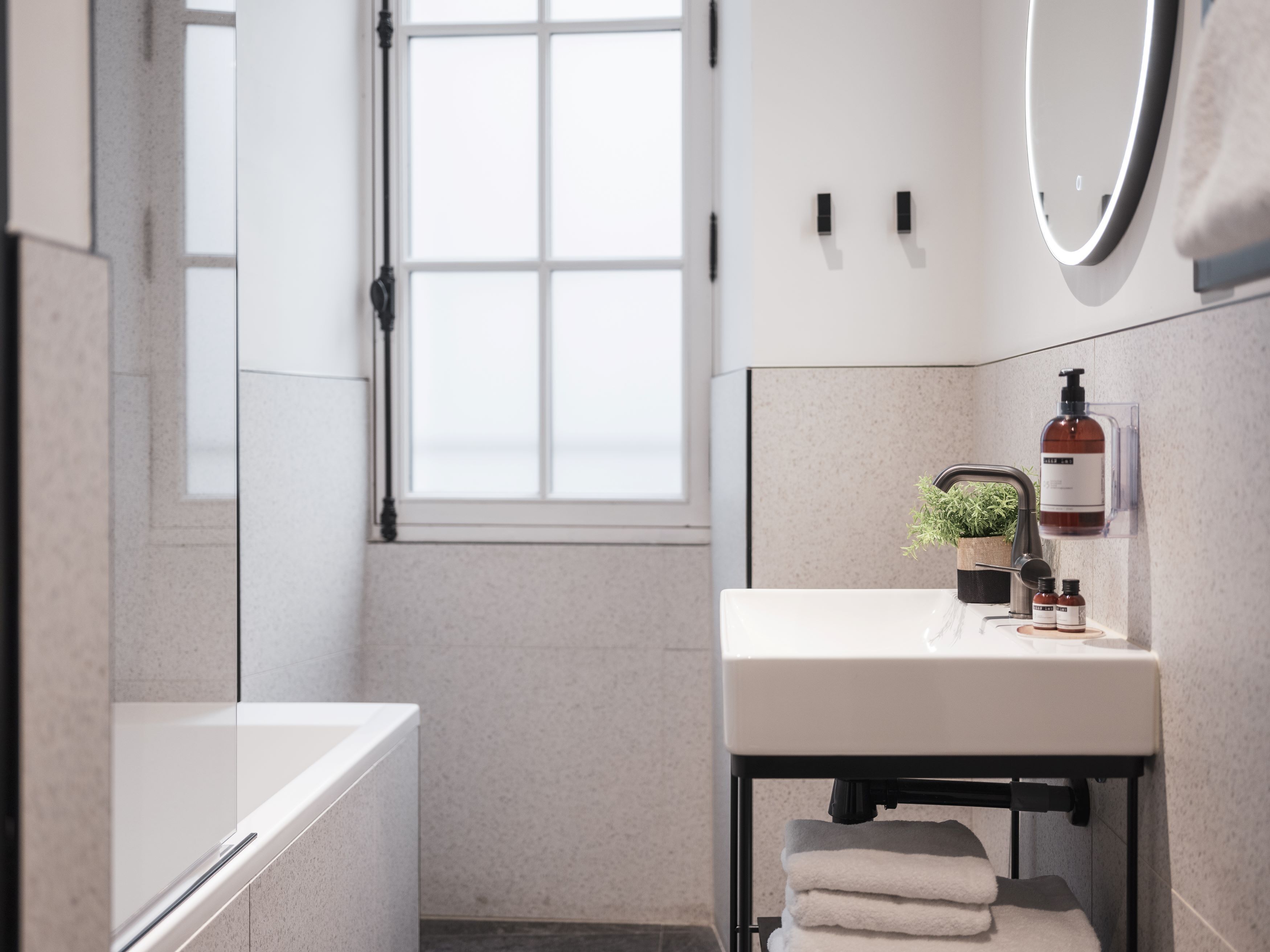 A modern bathroom with a white sink, a mirror, and a window. There are towels stacked under the sink and a small potted plant on the counter.