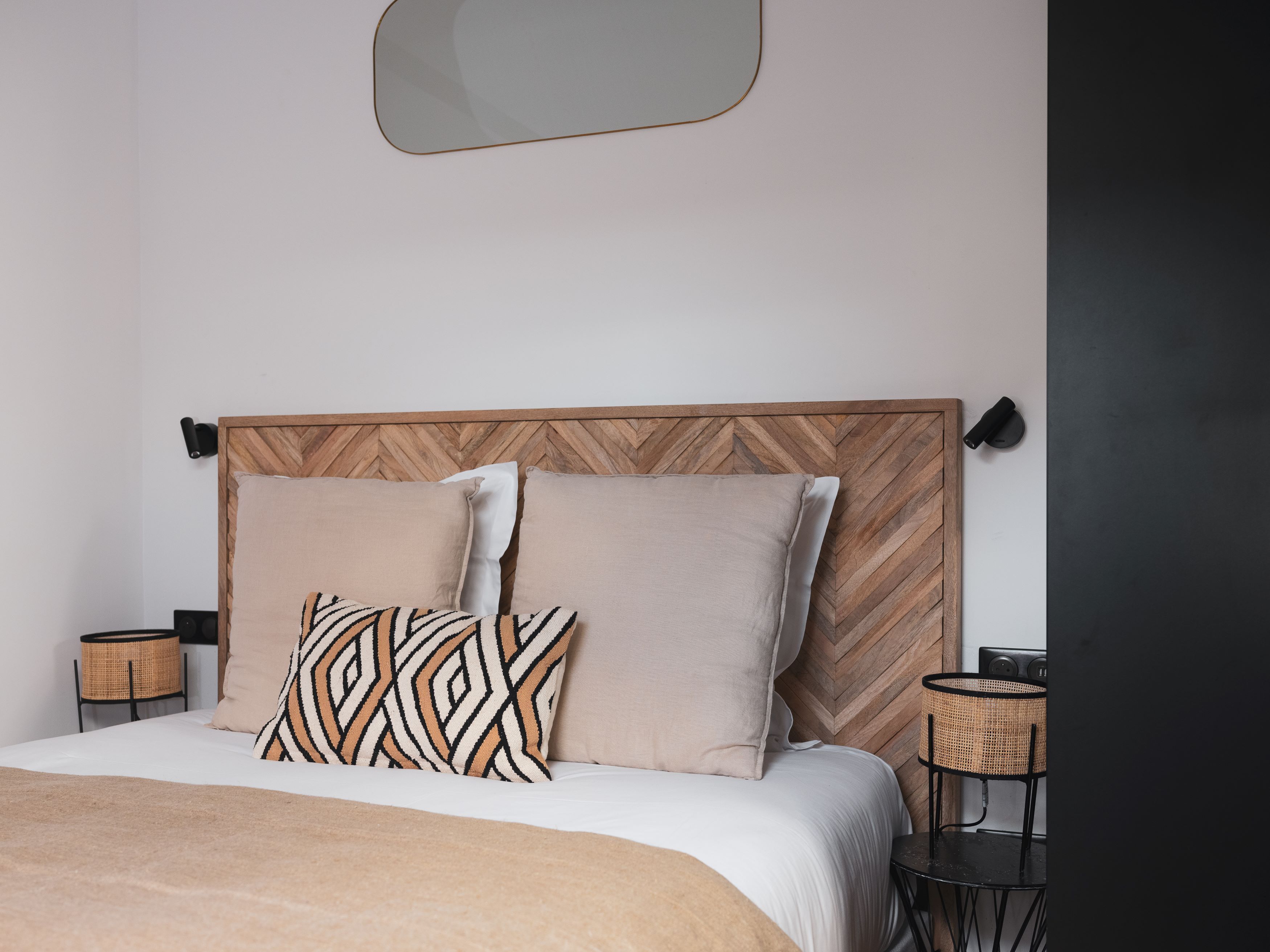 A modern bedroom with a herringbone-patterned headboard, two plain pillows, a patterned throw pillow, a bedside table with a lamp, and a small round table with a chair.