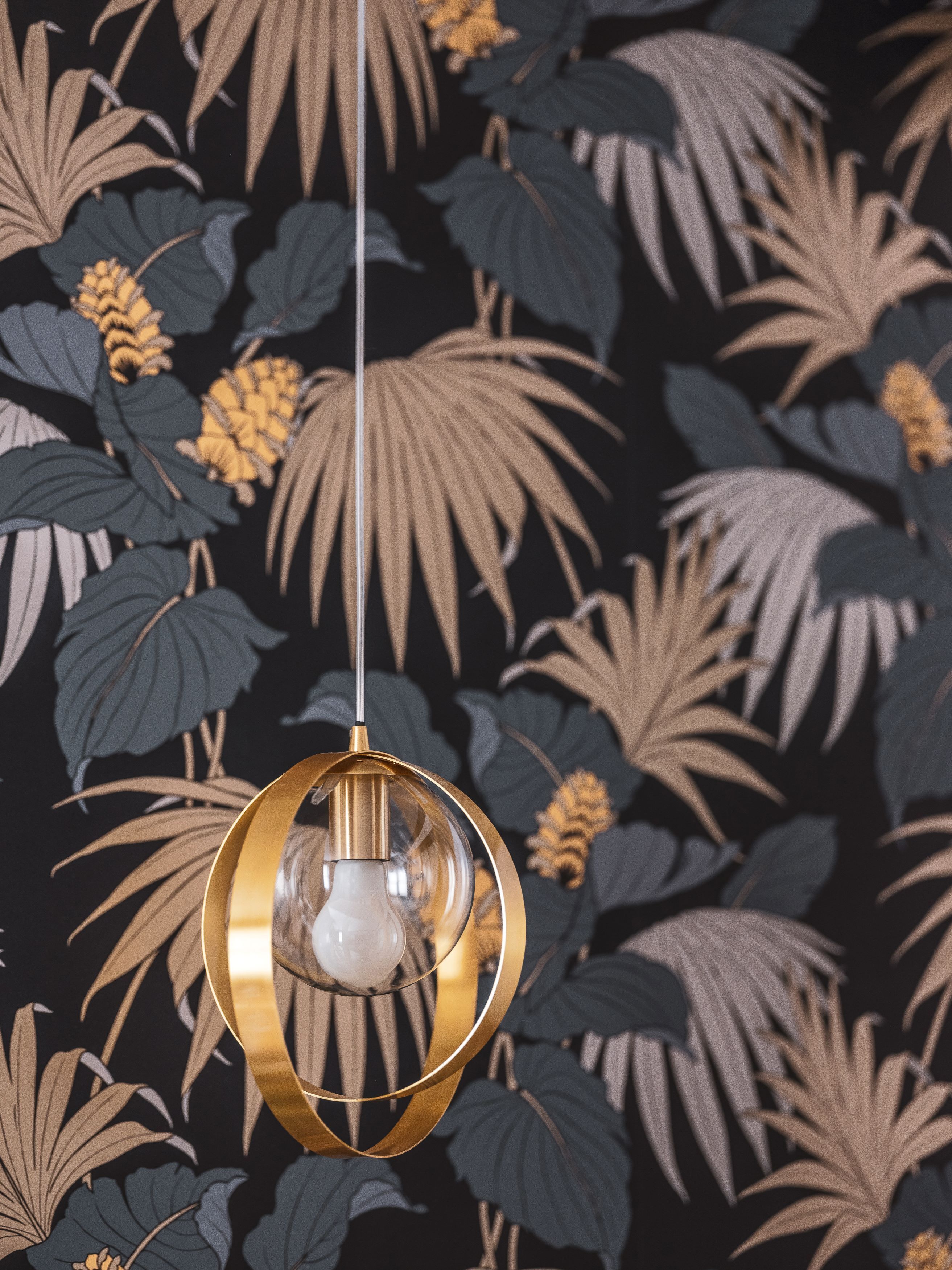 A hanging light fixture with a clear glass bulb and a gold-colored frame against a dark wall with a tropical leaf and flower pattern.