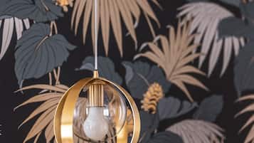 A hanging light fixture with a clear glass bulb and a gold-colored frame against a dark wall with a tropical leaf and flower pattern.