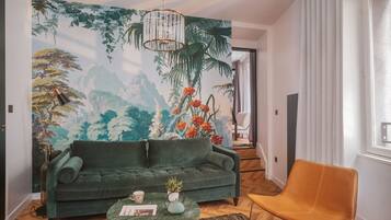 Uma sala de estar com um sofá verde, uma mesa de centro redonda, uma poltrona marrom, um mural com folhagem tropical e um lustre.