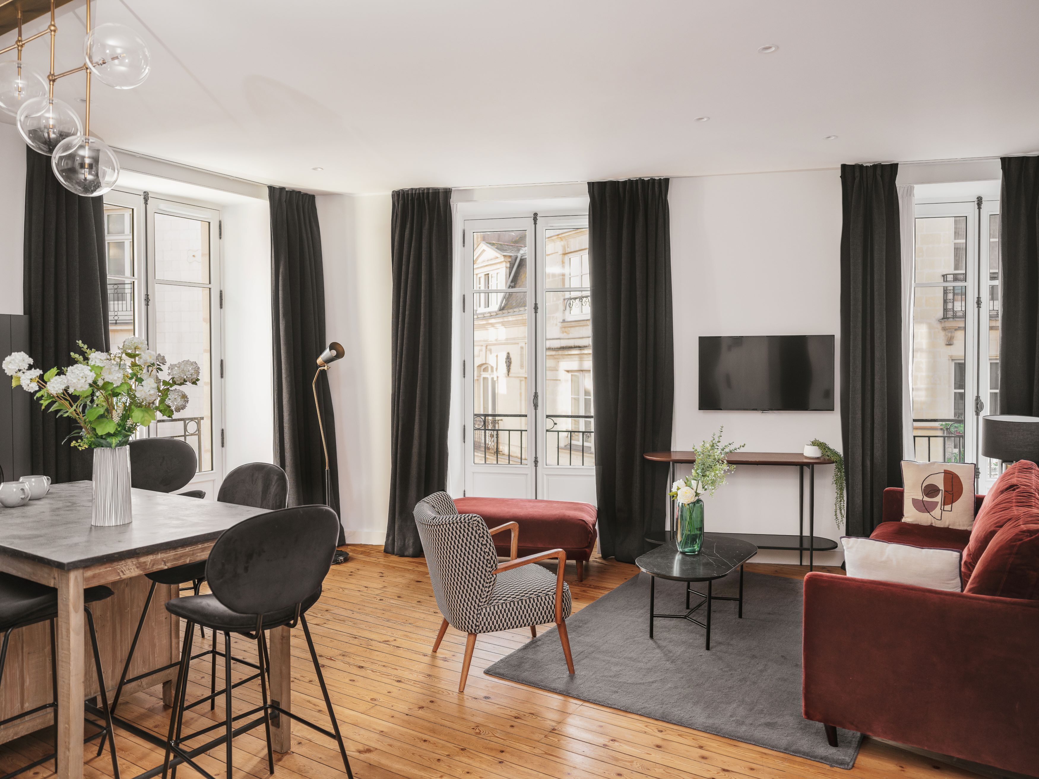 Una sala de estar moderna con piso de madera, un comedor con mesa blanca y sillas negras, un sofá rojo, un sillón estampado y un televisor montado en la pared.