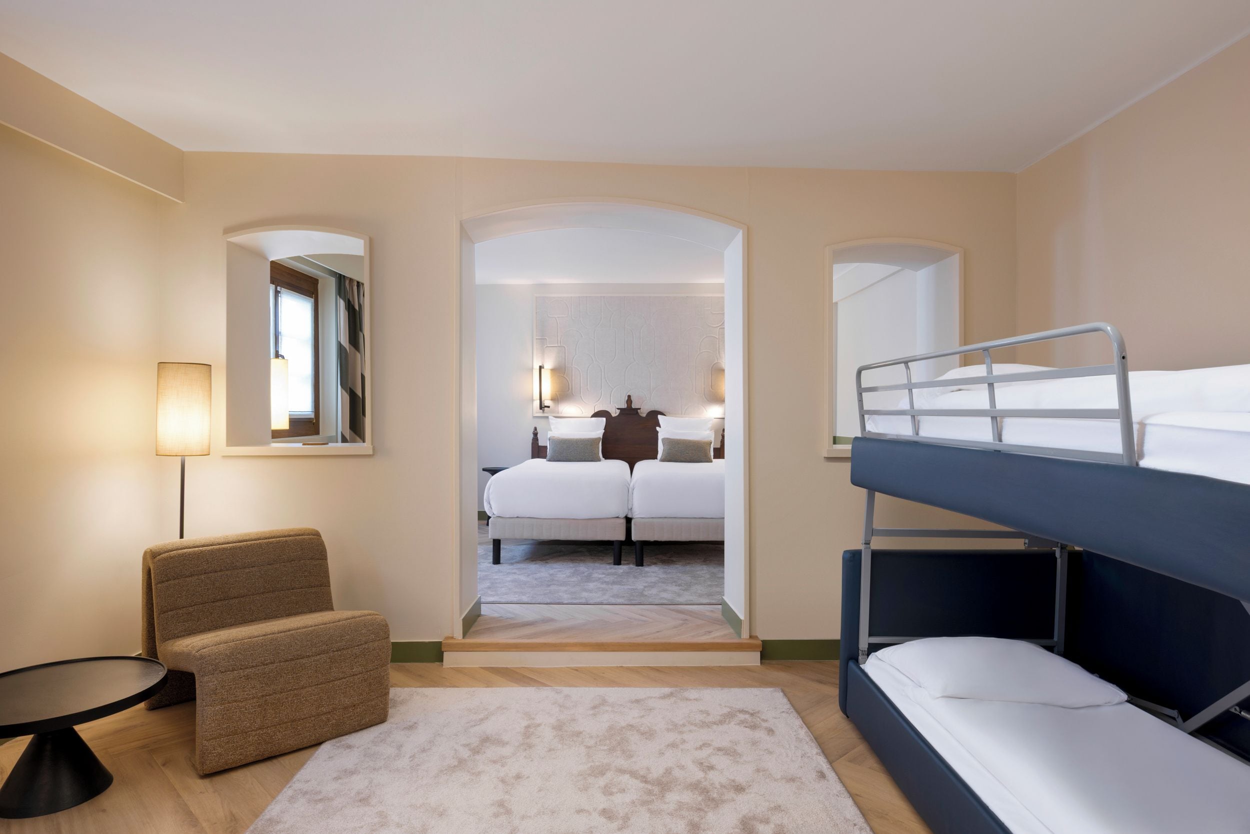 Ein Hotelzimmer mit Etagenbett, einem Einzelbett, einem Schreibtisch mit Stuhl, einer Lampe und Blick auf den Flur.