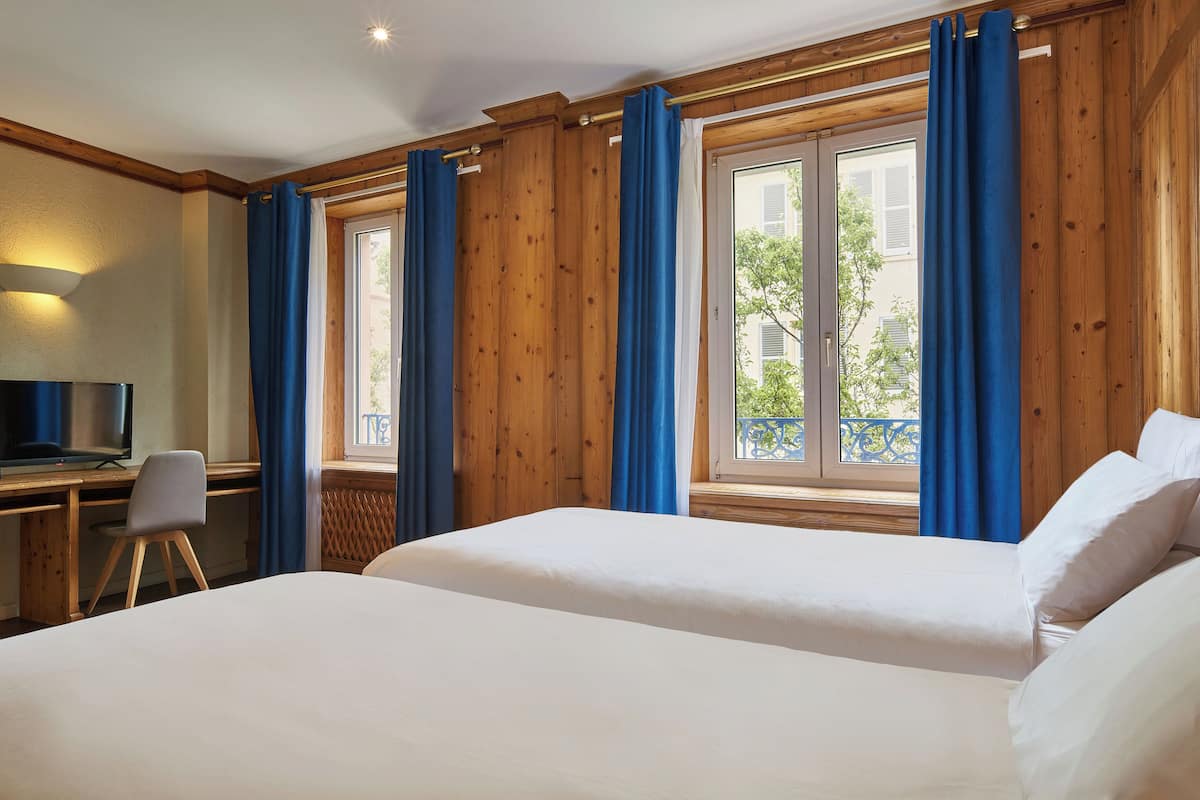 A hotel room with two beds, a desk with a chair, and large windows with blue curtains.