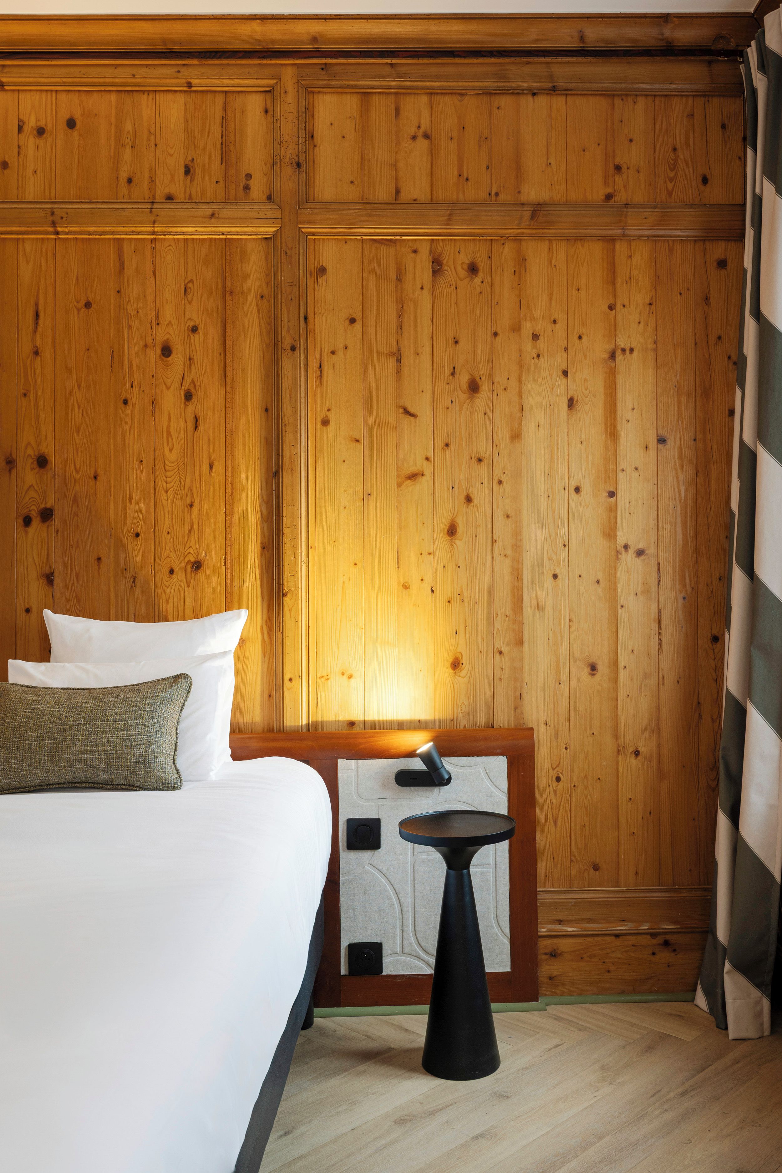 Quarto de hotel com parede revestida de madeira, cama com roupa de cama branca, uma mesinha e um criado-mudo.