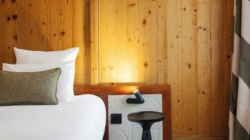 Quarto de hotel com parede revestida de madeira, cama com roupa de cama branca, uma mesinha e um criado-mudo.