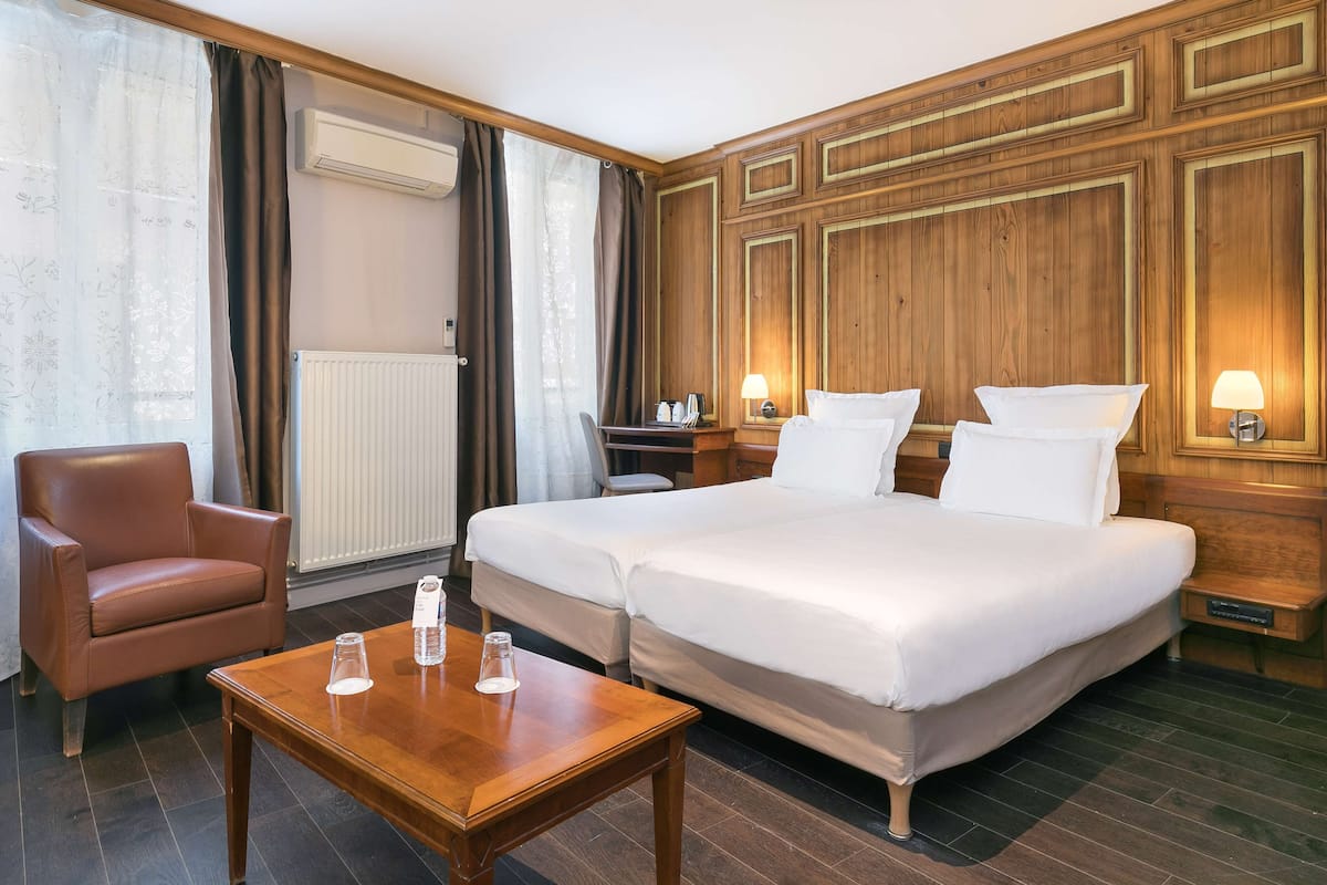 A hotel room with two beds, a wooden headboard, a wooden table, a leather chair, and a window with curtains.
