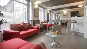 Una hall moderna di un hotel con divani rossi, una sala da pranzo con tavolo in vetro e una cucina sullo sfondo.