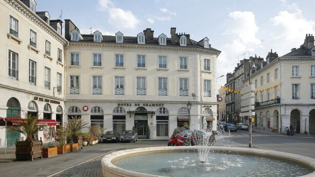 Un edificio storico con una fontana al centro, circondato da strade acciottolate e auto parcheggiate.