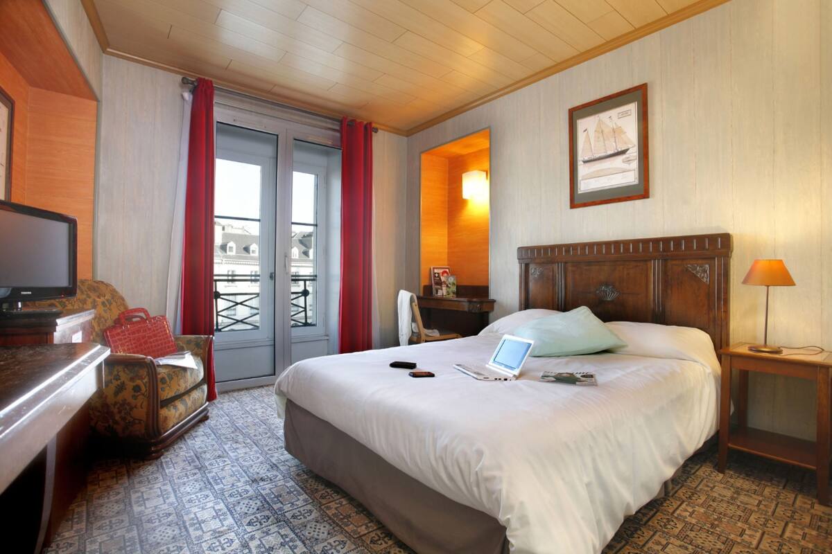 A hotel room with a bed, a desk with a laptop, a chair, a TV, and a window with curtains.
