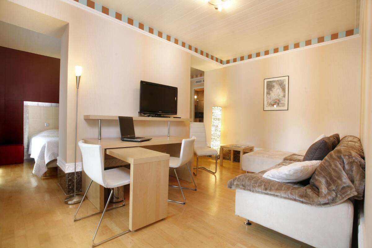 A modern hotel room with a wooden desk, a flat-screen TV, a sofa, and a bed.