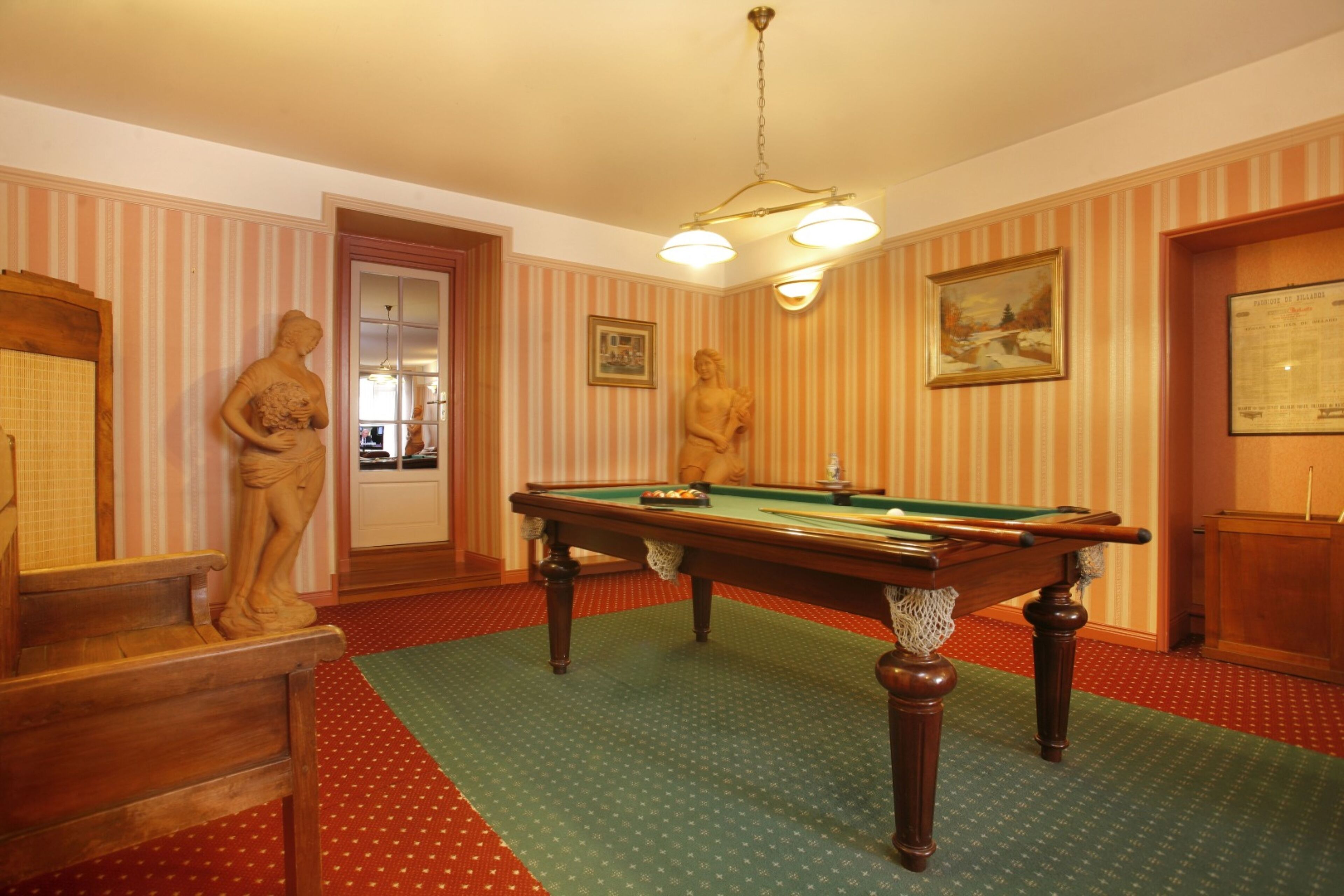 Una sala de billar con papel tapiz a rayas, una mesa de billar y dos estatuas.