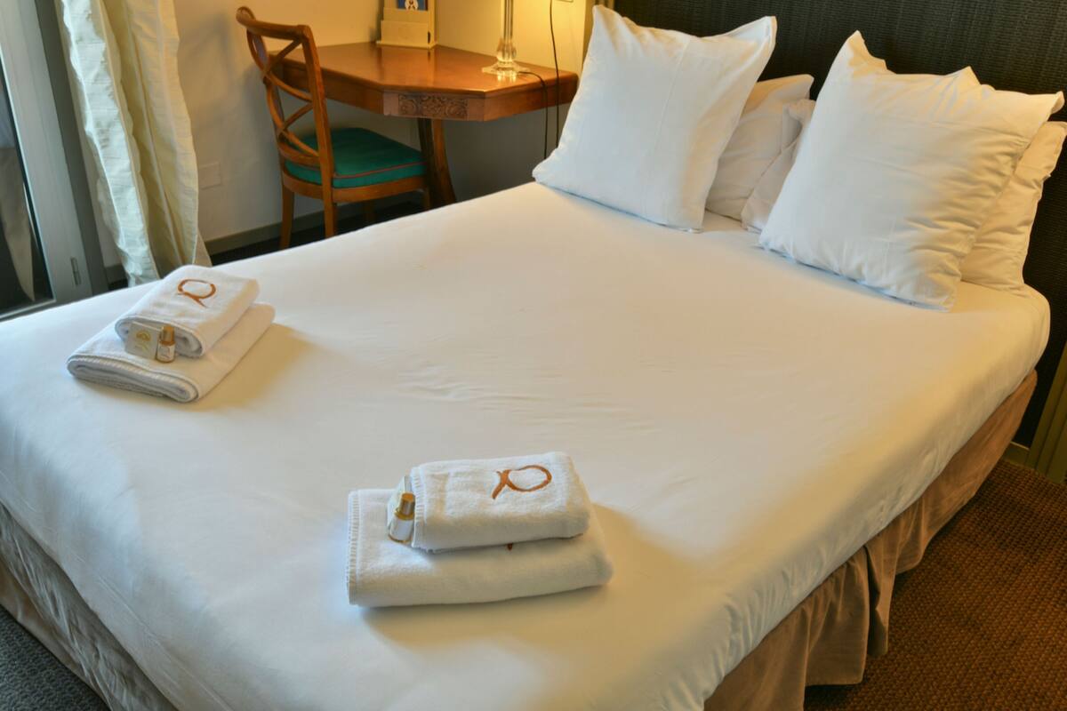 A neatly made bed with white linens and two towels with a monogrammed towel ring.