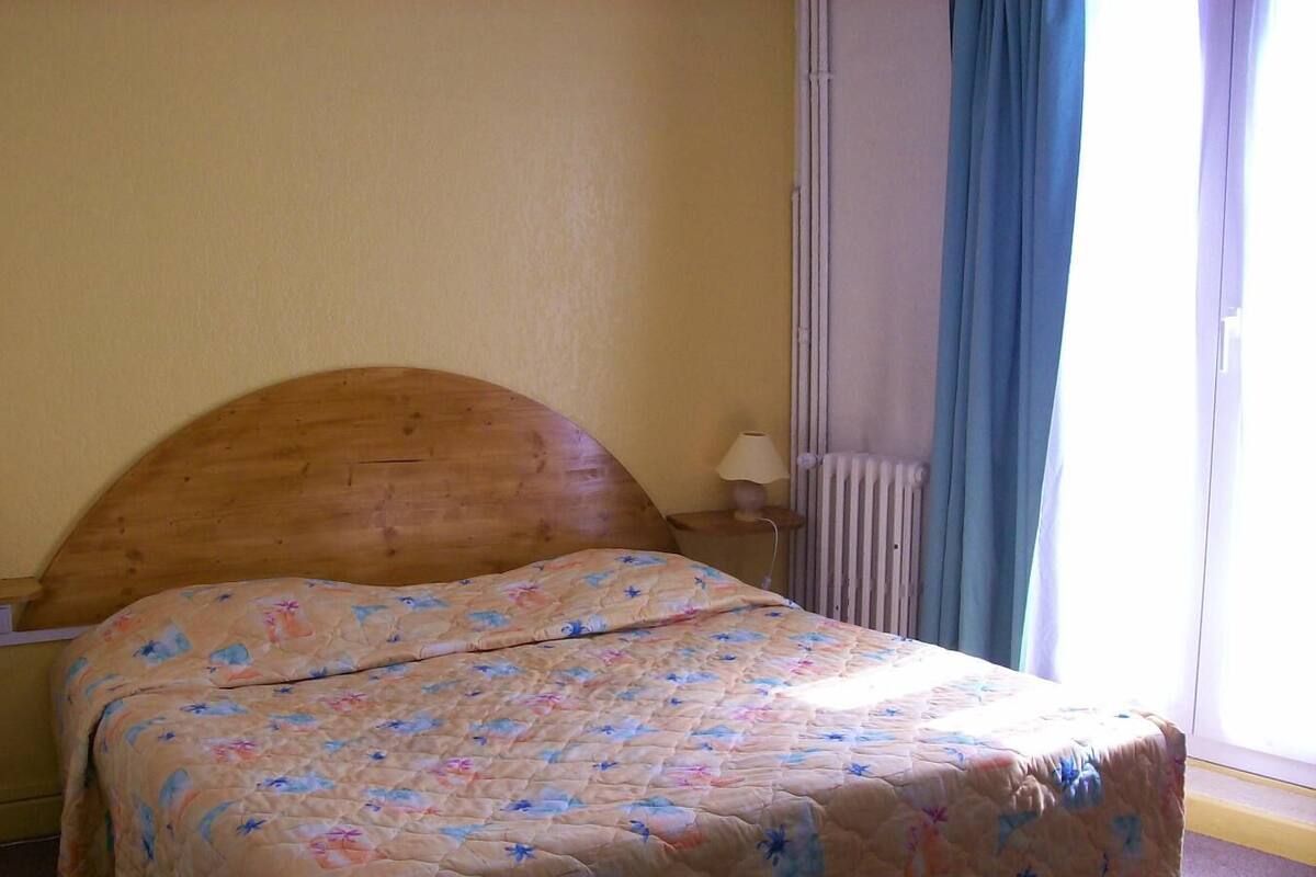 A single bed with a wooden headboard in a room with a plain yellow wall and a window with blue curtains.
