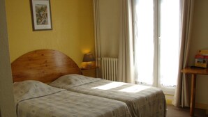Une chambre avec deux lits, une tête de lit en bois, une table de chevet avec une lampe, un radiateur et une fenêtre avec des rideaux.