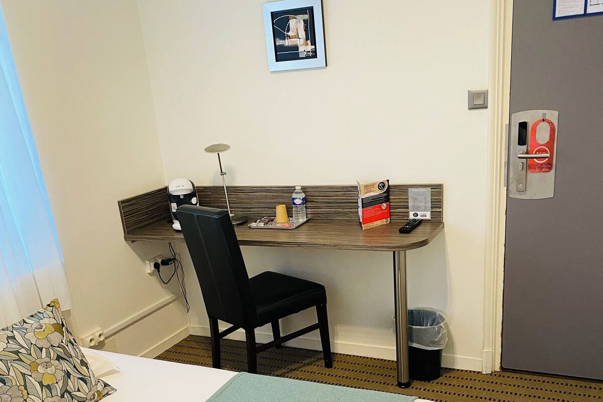 Une chambre d’hôtel avec un lit, un bureau et une chaise, un tableau au mur et une poubelle.