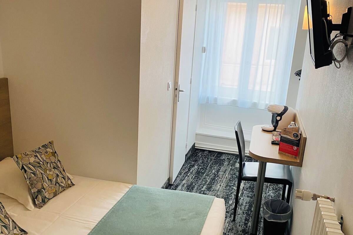 Une chambre d’hôtel avec un lit, un bureau, une chaise, un radiateur et une fenêtre avec des rideaux.