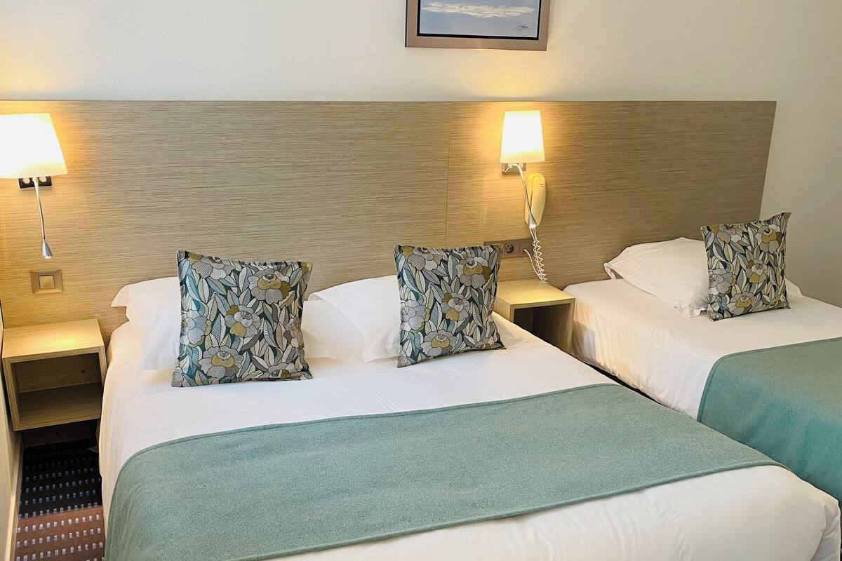 Une chambre d’hôtel avec deux lits, chacun recouvert d’une couverture verte et agrémenté d’oreillers à motifs floraux, une tête de lit en bois, des lampes de chevet et un tableau encadré au mur.