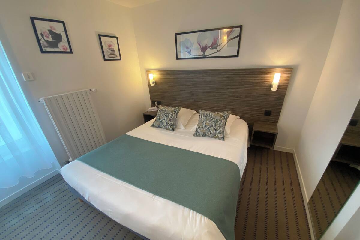 Une chambre d’hôtel avec un grand lit, une tête de lit dotée d’un éclairage intégré, un radiateur et deux œuvres d’art florales encadrées accrochées au mur.