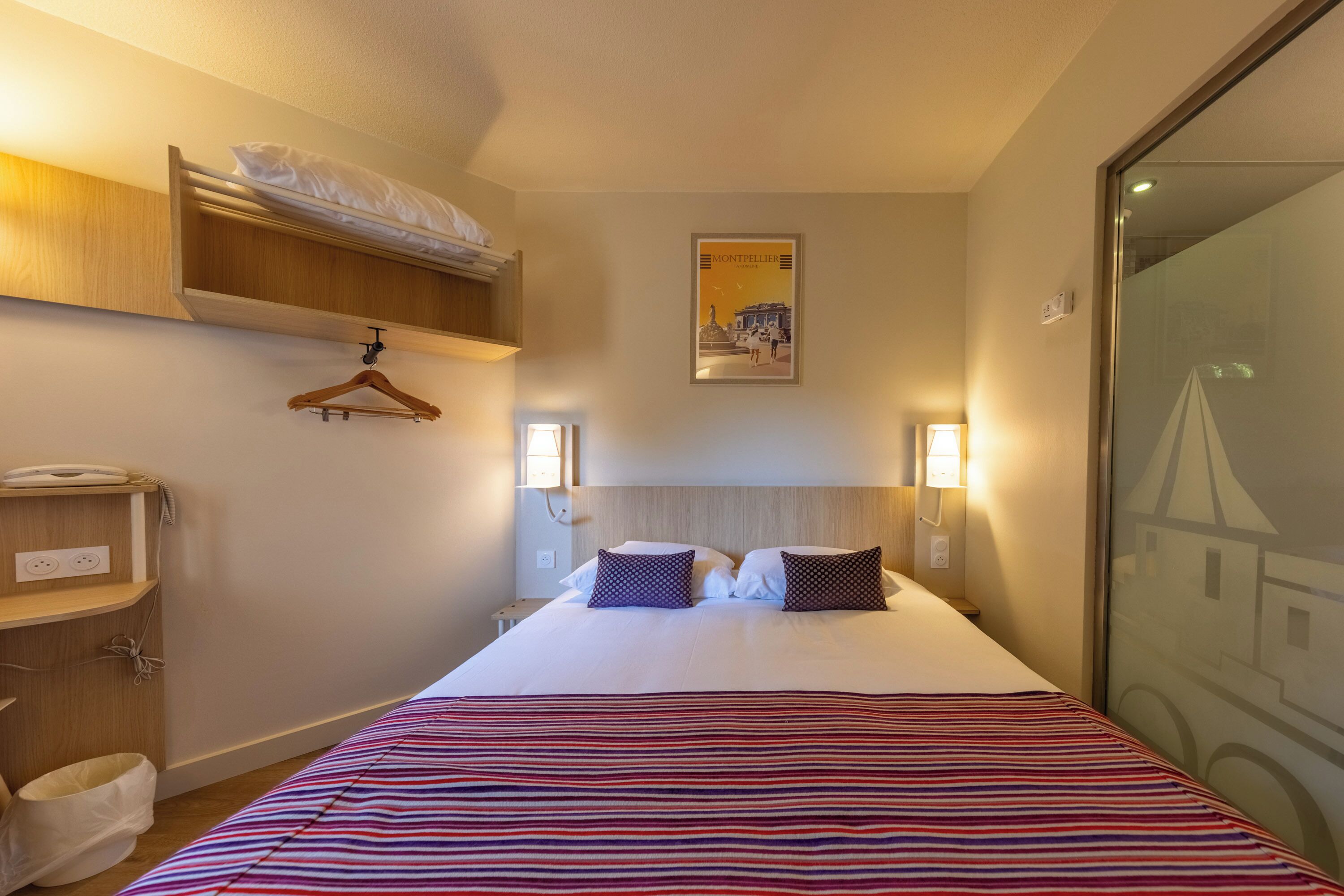 Une chambre d’hôtel avec un lit, une armoire suspendue, une table de chevet, une lampe et une affiche au mur.