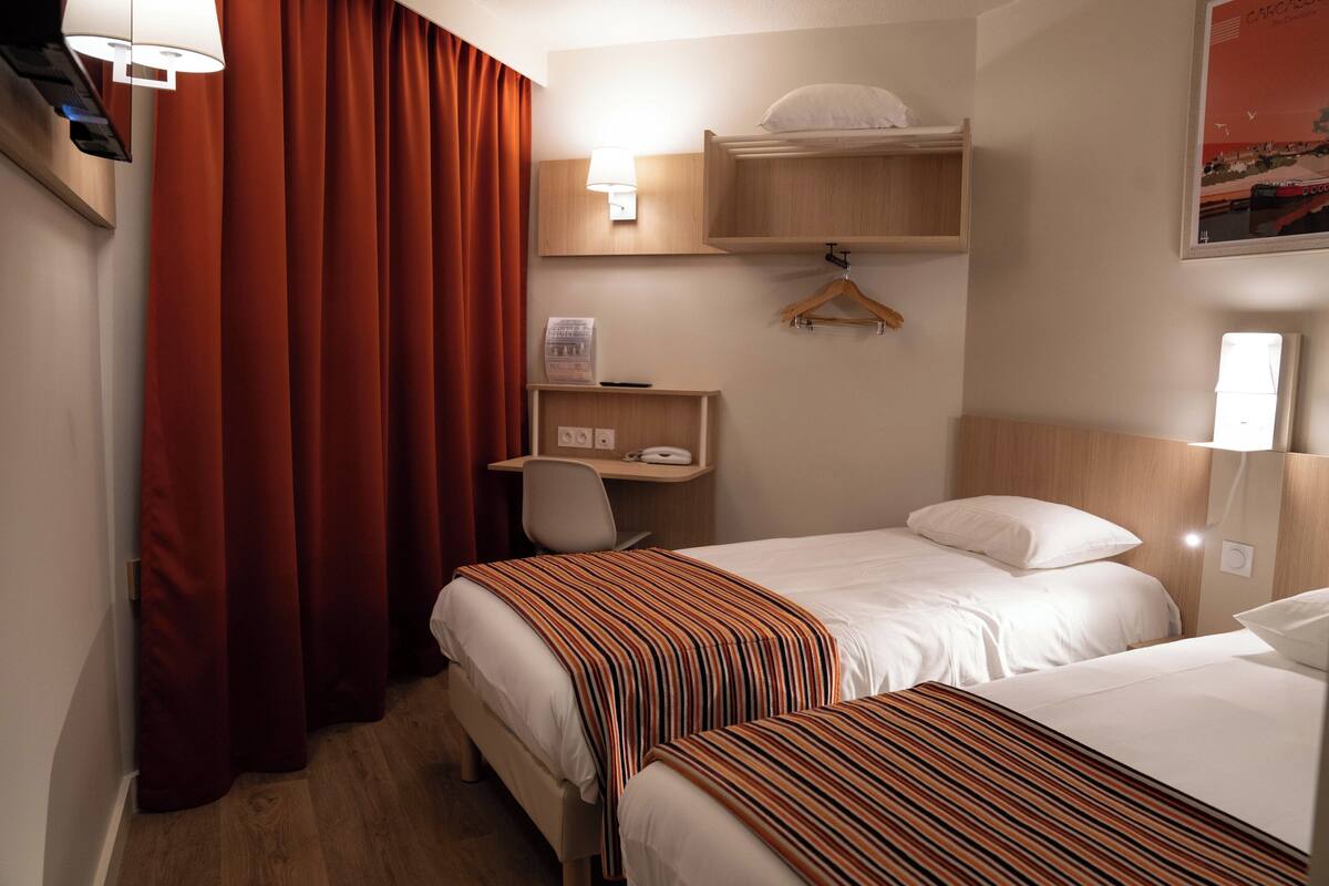 Habitación de hotel con dos camas, un escritorio, una silla, perchero y estante en la pared.
