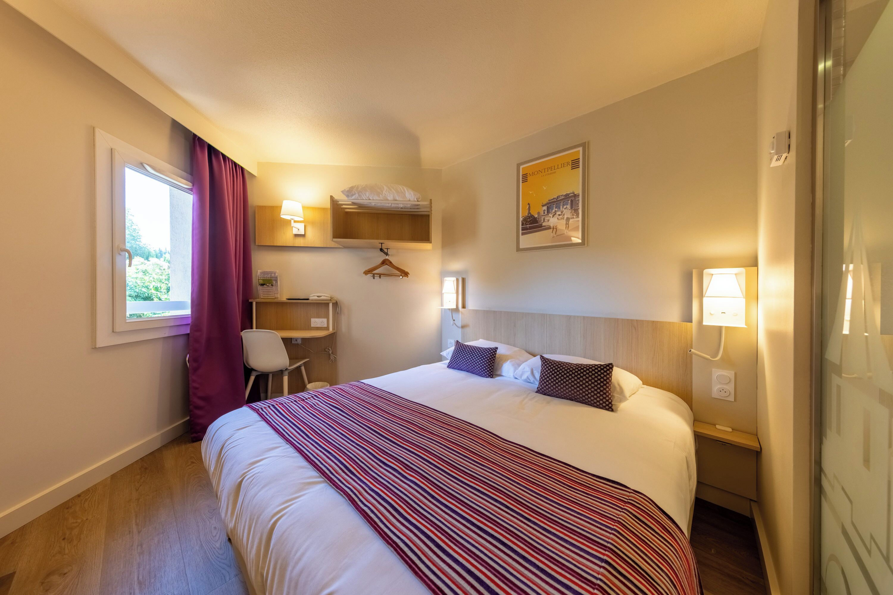 A hotel room with a bed, a desk, a chair, a window with curtains, a wall-mounted lamp, and a poster.