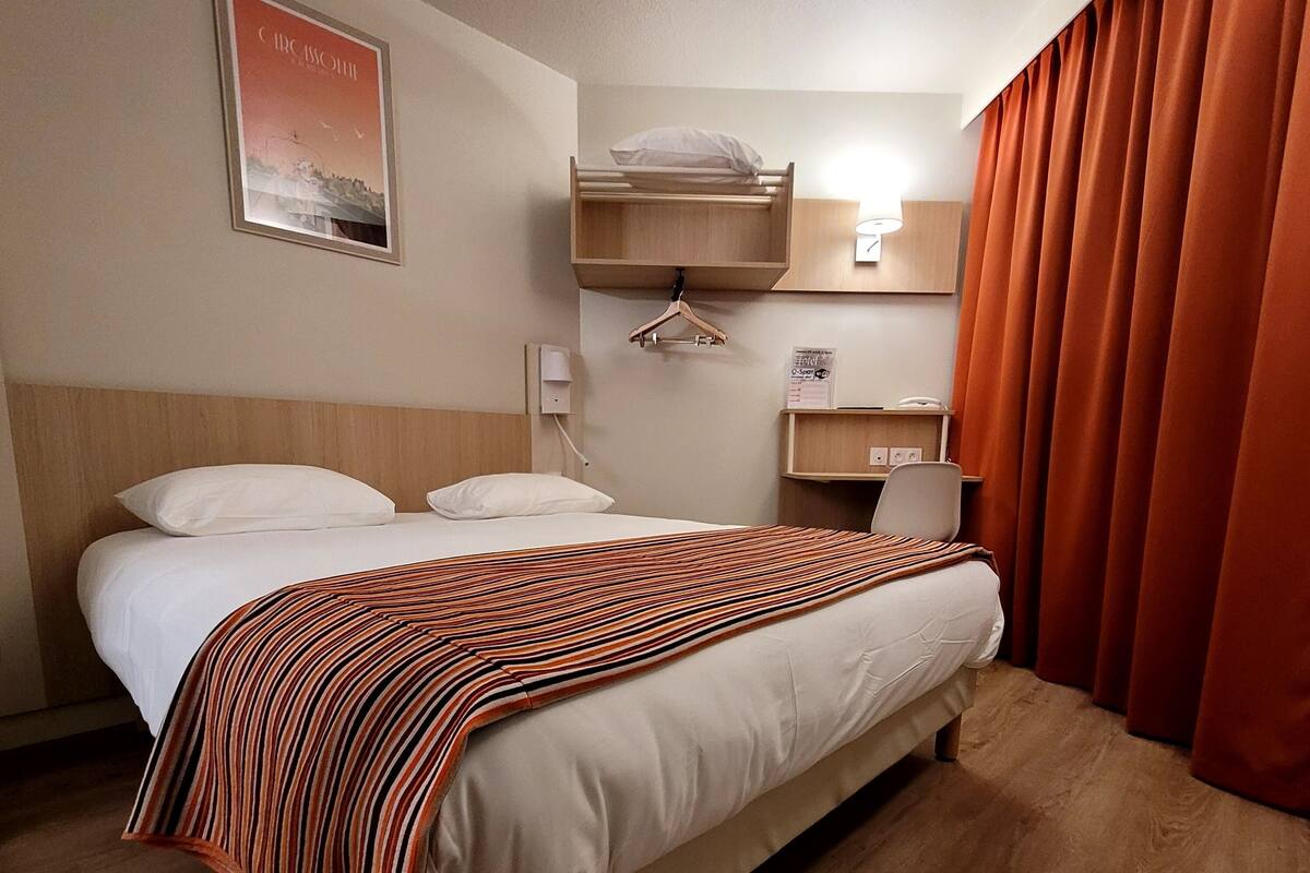 Habitación de hotel con cama, cabecera de madera, perchero colgante, mesita de noche con lámpara y un cartel que indica los servicios del hotel.