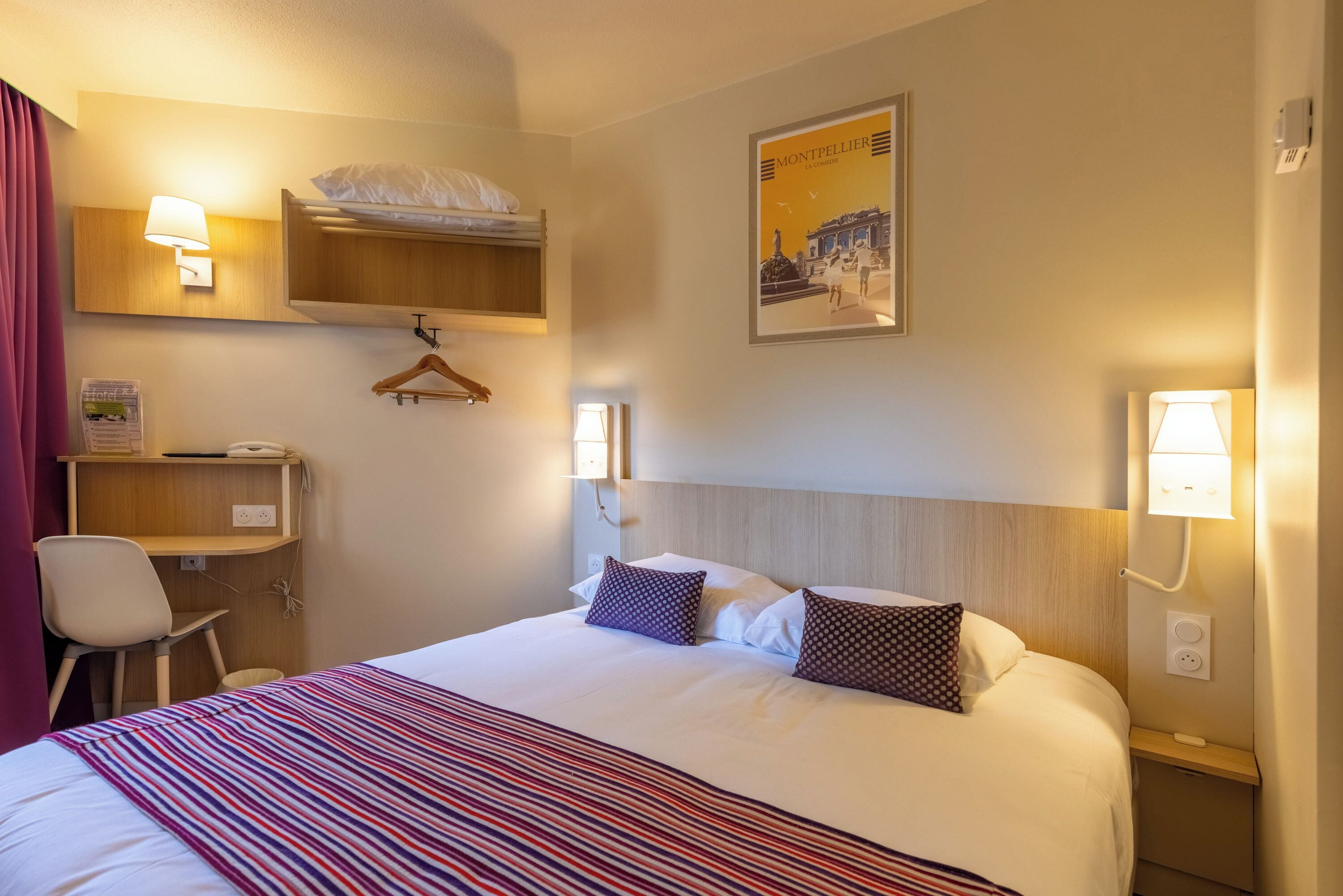 A hotel room with a bed, a desk, a chair, a hanging coat rack, and a wall-mounted lamp.