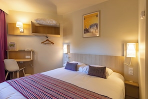 Habitación de hotel con cama, escritorio, silla, perchero y lámpara de pared.
