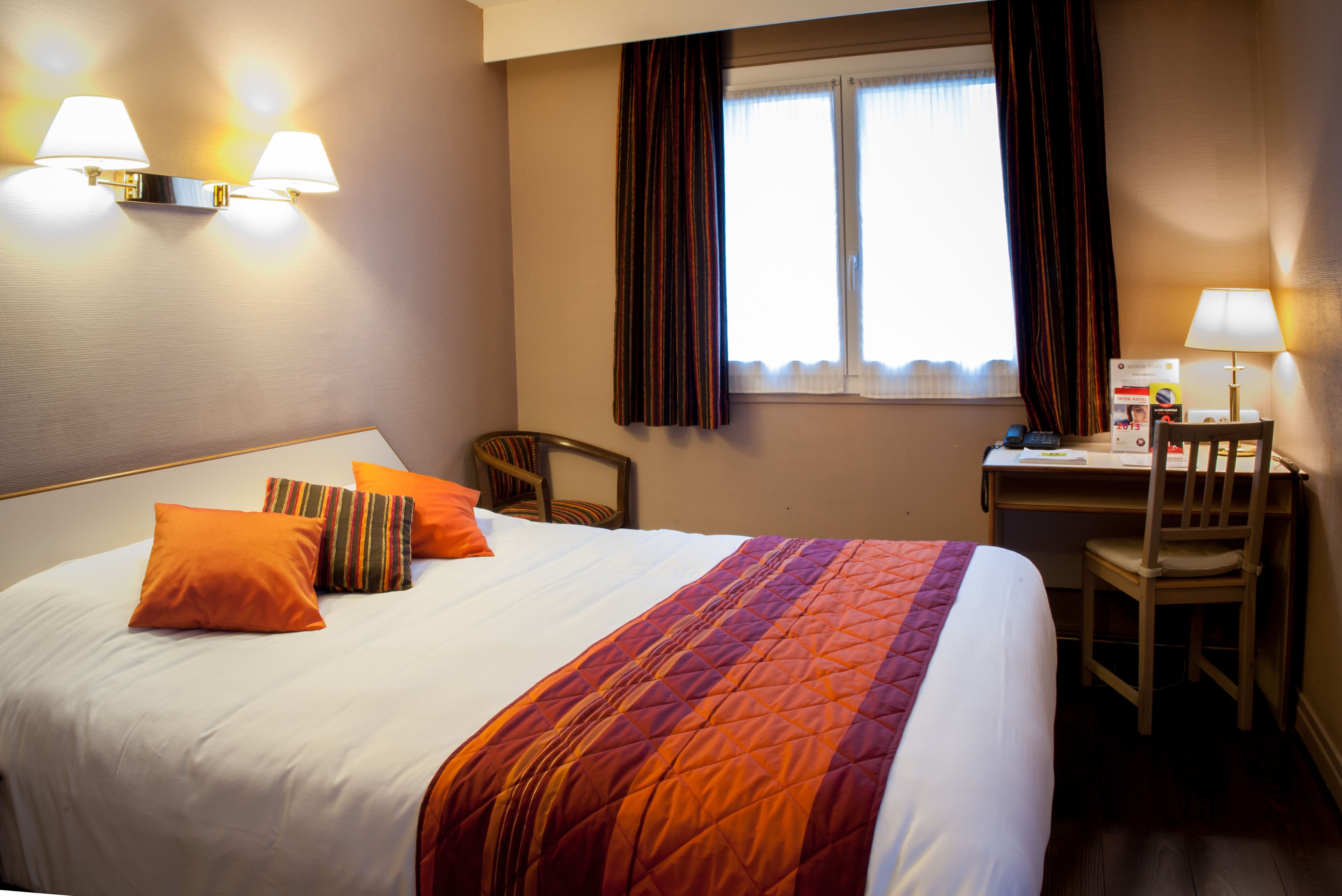 A hotel room with a large bed, two pillows, a desk with a chair, and a window with curtains.