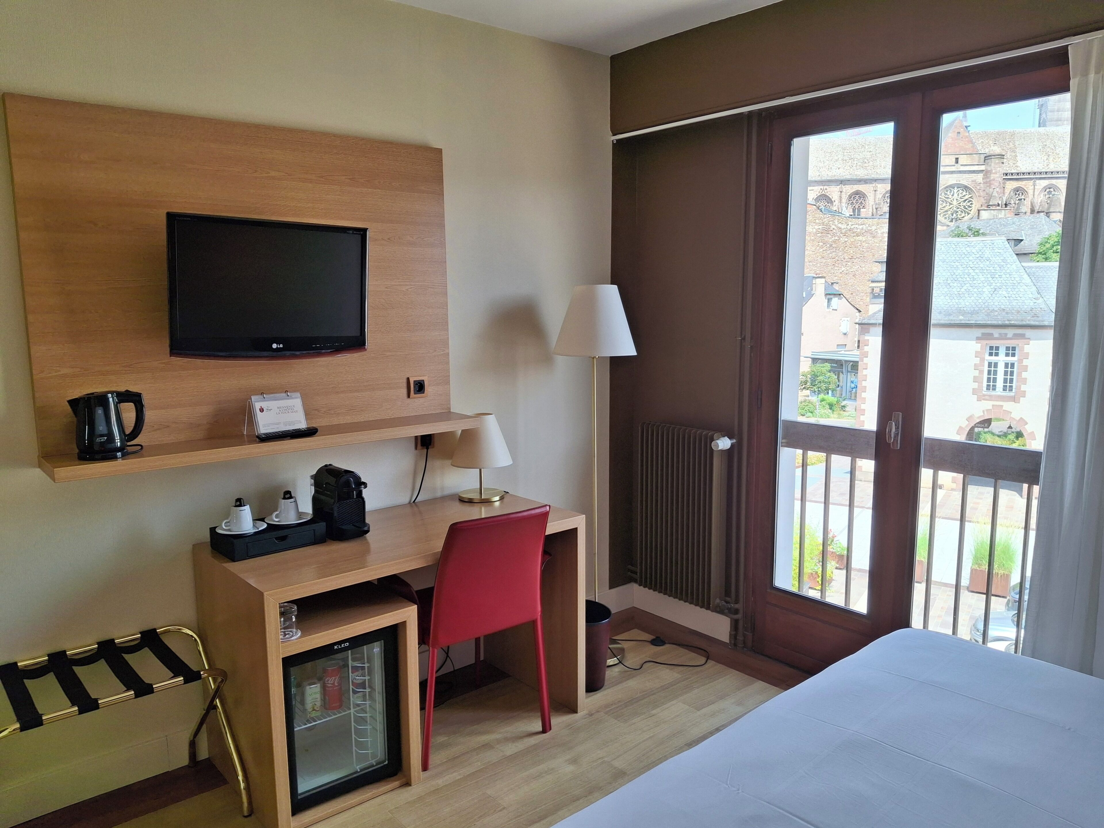 A hotel room with a bed, a desk with a chair, a television, a coffee maker, a mini-fridge, and a window with a view of buildings.