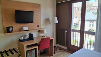 A hotel room with a bed, a desk with a chair, a television, a coffee maker, a mini-fridge, and a window with a view of buildings.