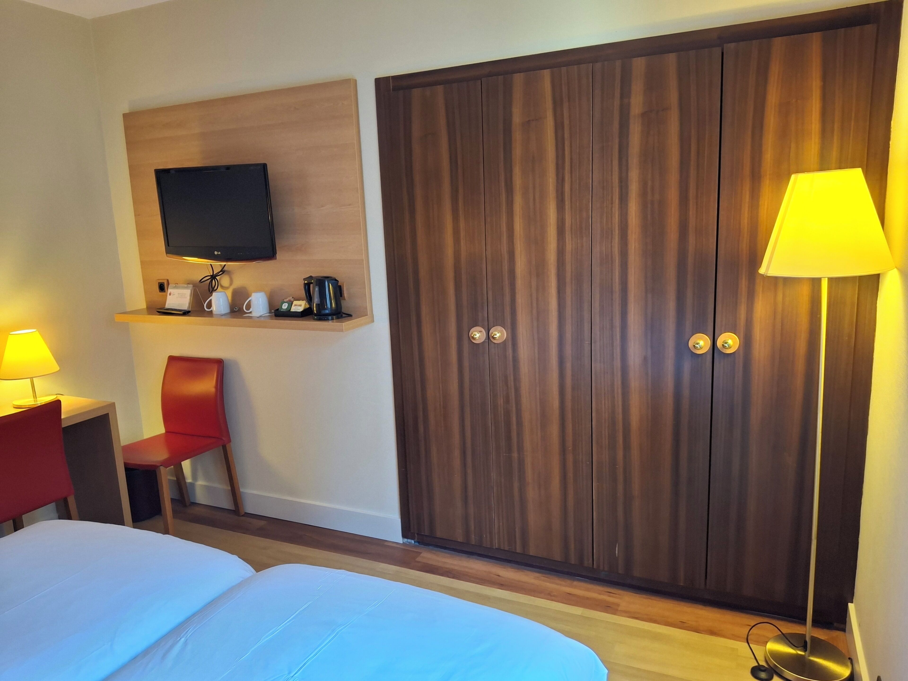 Una habitaci ón de hotel con cama, escritorio con lámpara, televisor montado en la pared, armario y mesita de noche con lámpara.