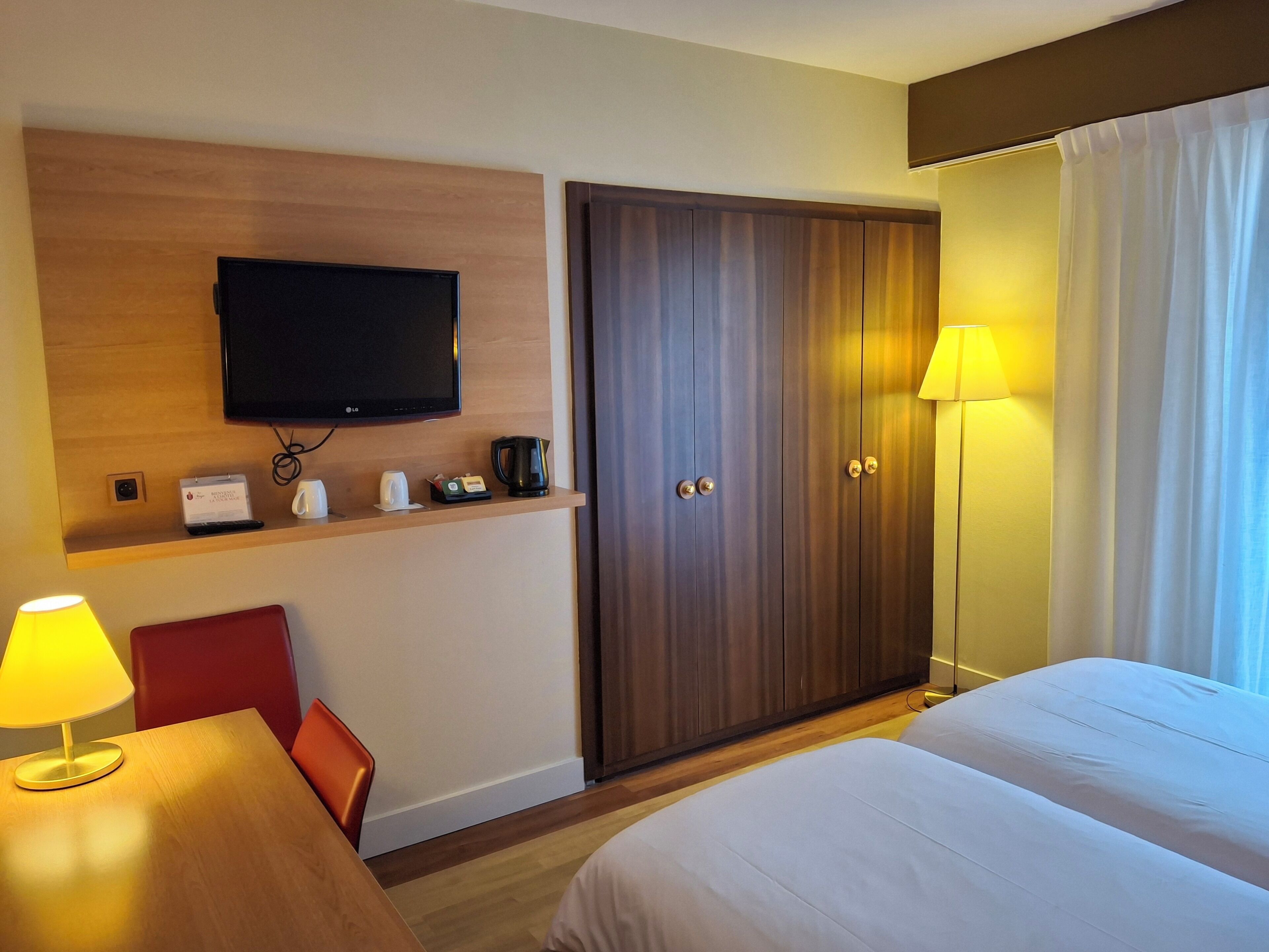 A hotel room with a bed, a desk with a lamp, a television mounted on the wall, a wardrobe, and a bedside table with a lamp.