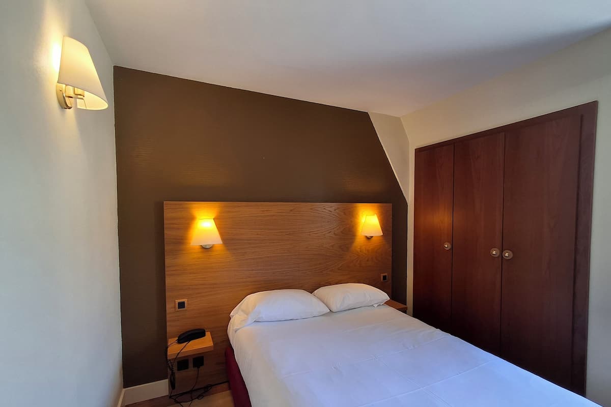 A bedroom with a wooden headboard, two bedside lamps, a bed with white linens, a wooden nightstand with a phone, and a wardrobe.